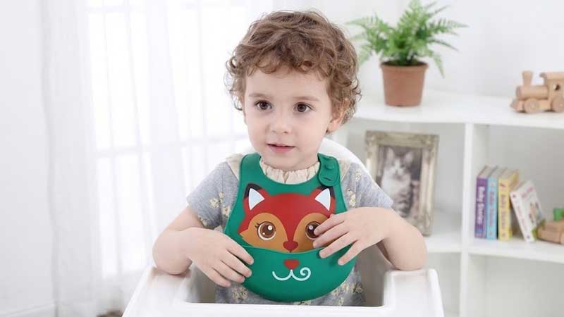 लोमड़ी के आकार की सिलिकॉन बिब पहने एक प्यारा सा घुंघराले बालों वाला बच्चा